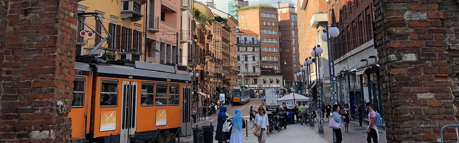A street with many different colored buildings, including several red brick buildings. Two orange cable-car trains are passing by in opposite directions in the middle of the street. 